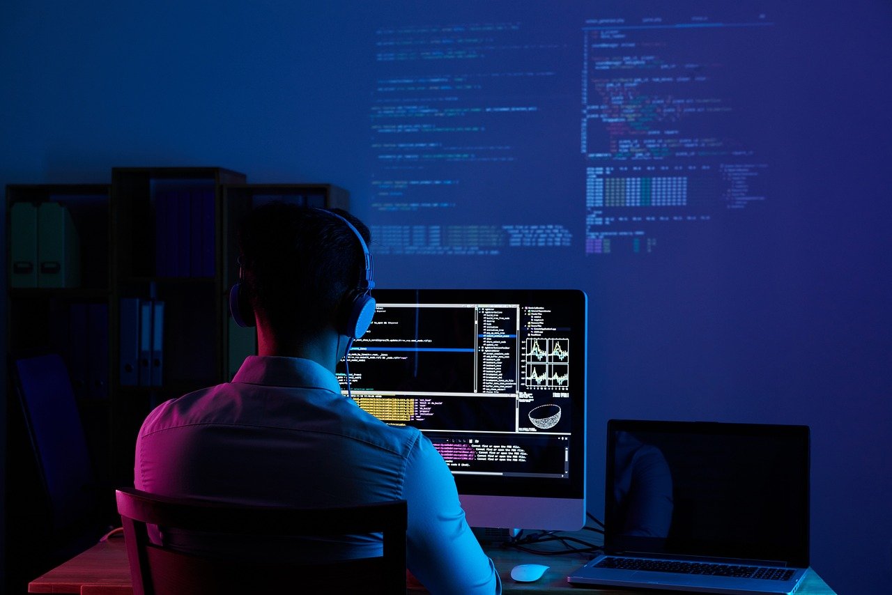A software developer wearing headphones working on multiple monitors with code and data visualizations projected on the wall in a dark room.